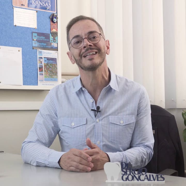O professor sorri, sentado à mesa da sala dele na UNESP; no mural, uma flâmula da University of Kansas