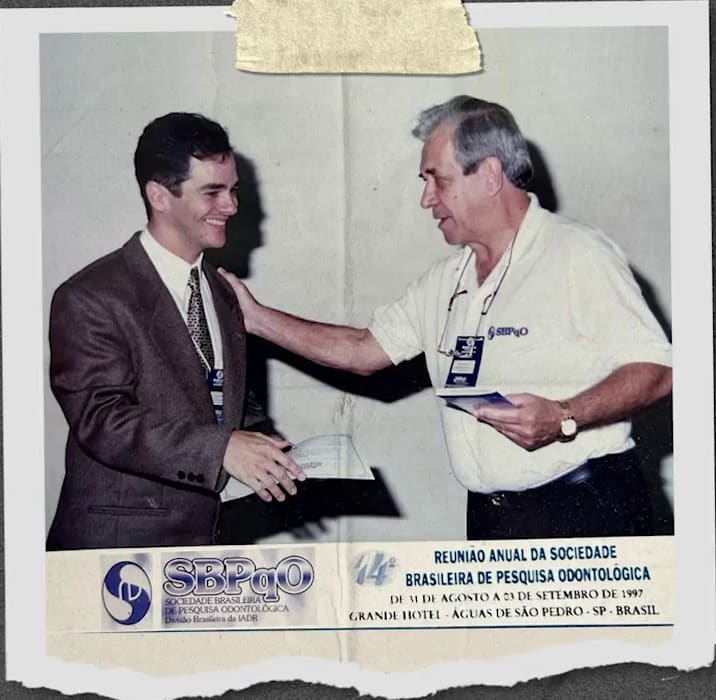 Recorte de jornal de 1997, preso com fita: o jovem Sérgio Gonçalves, de terno, cumprimentado ao receber um certificado na 14ª Reunião Anual da SBPqO